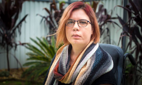 Image of Kelly Cox, Kelly has light brown hair, is wearing glasses, and a colourful scarf. There are plants that are blurry in the background.