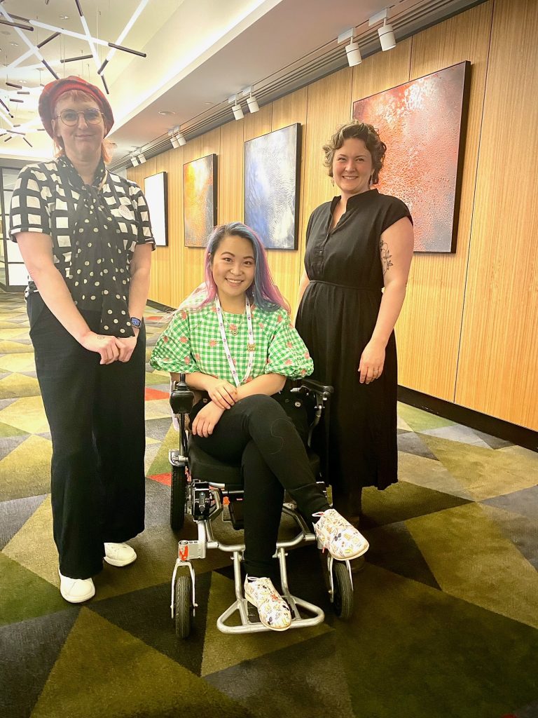 Photo of three people smiling at the camera. Saph has a red hat and black and white top, Akii has pink and blue hair and is wearing a green top sitting in a wheelchair and Zoe is wearing a black dress with short curly hair.