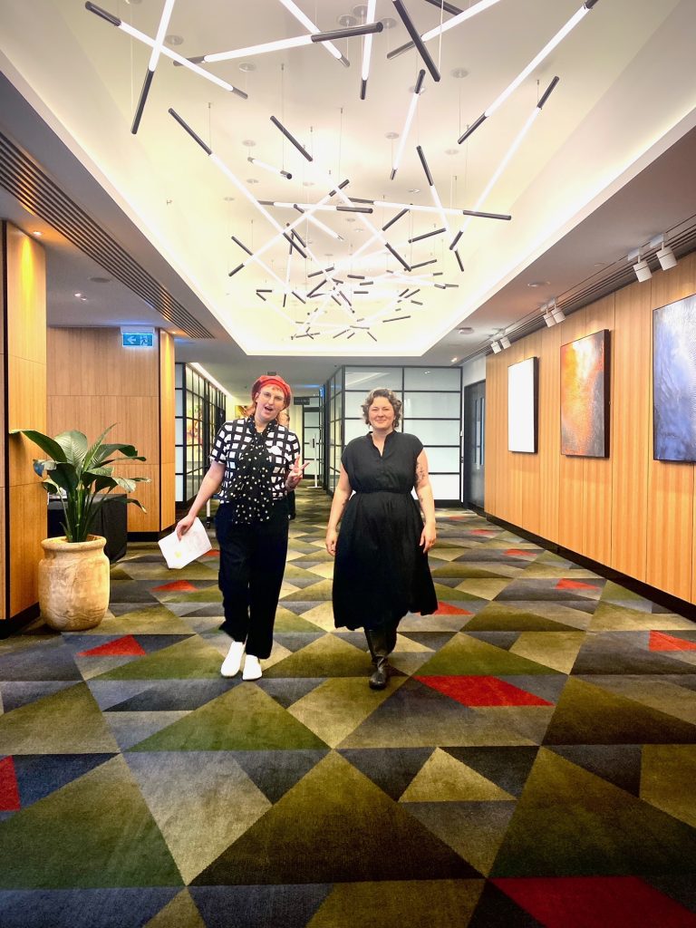 Photo of two people smiling at the camera. Saph has a red hat and black and white top and Zoe is wearing a black dress with short curly hair.