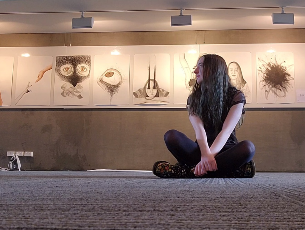 [Image: Young girl wearing all black sitting cross-legged with an exhibit of her art behind her.]