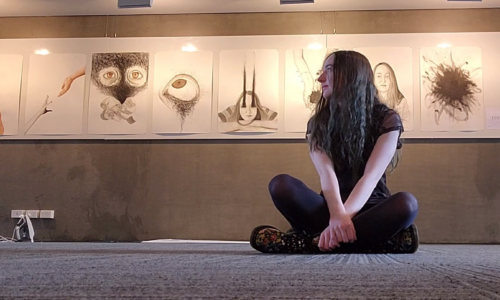 [Image: Young girl wearing all black sitting cross-legged with an exhibit of her art behind her.]