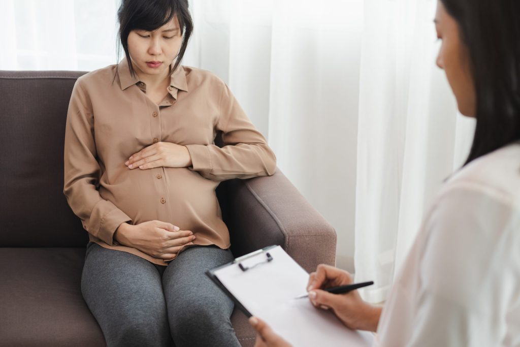 Photo of a women sitting on a couch opposite a professional in a white coat and clipboard. The woman is pregnant hugging her belly.