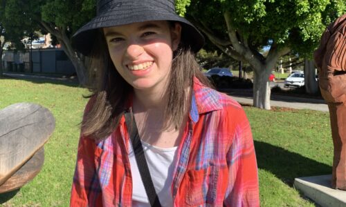 A photo of Nicola, a woman in her twenties, smiling with medium length brown hair and brown eyes. Nicola is wearing a black hat and skirt, and a white shirt with a red and blue checked button up over the top, tied at her waist. Nicola is standing in a park, with green grass and big, leafy trees in the background behind her.