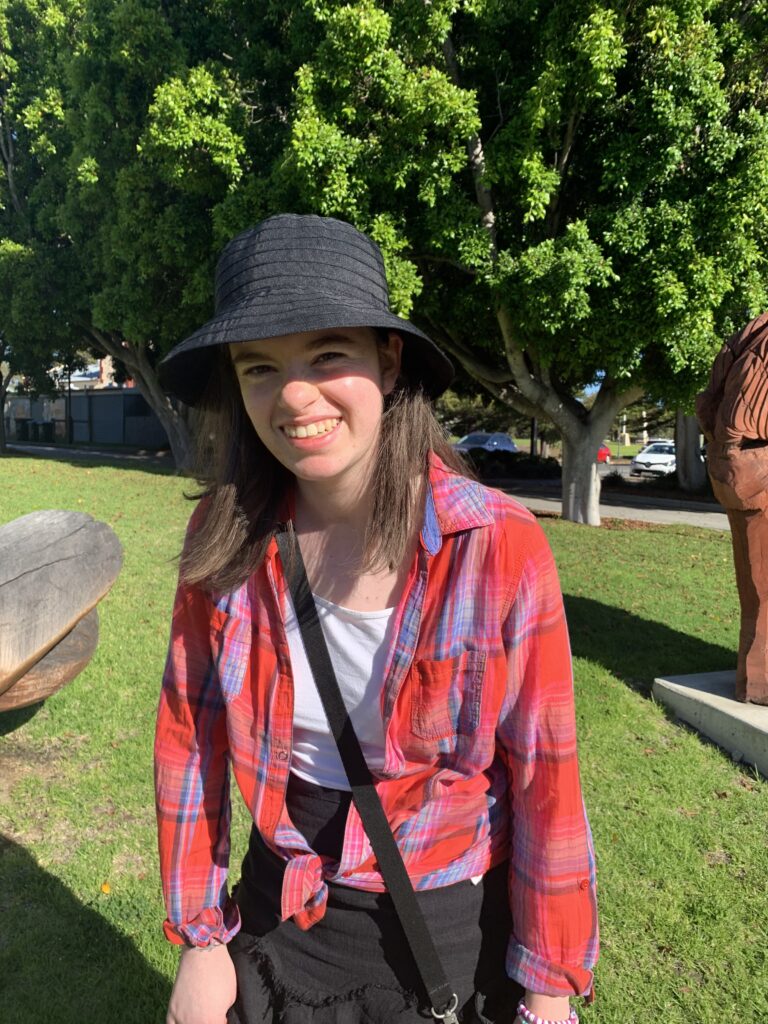 A photo of Nicola, a woman in her twenties, smiling with medium length brown hair and brown eyes. Nicola is wearing a black hat and skirt, and a white shirt with a red and blue checked button up over the top, tied at her waist. Nicola is standing in a park, with green grass and big, leafy trees in the background behind her.