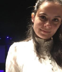 A mixed-race, medium-skinned woman in her mid-twenties. She has dark brown hair in a half-up half-down style, and is wearing a white button-up shirt.