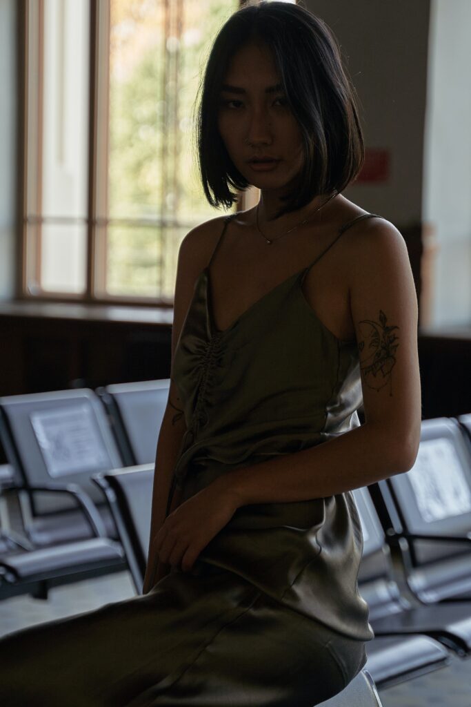 Woman in a grey dress leans on a seat in a darkened hospital waiting room. Her expression is deadpan and forlorn.