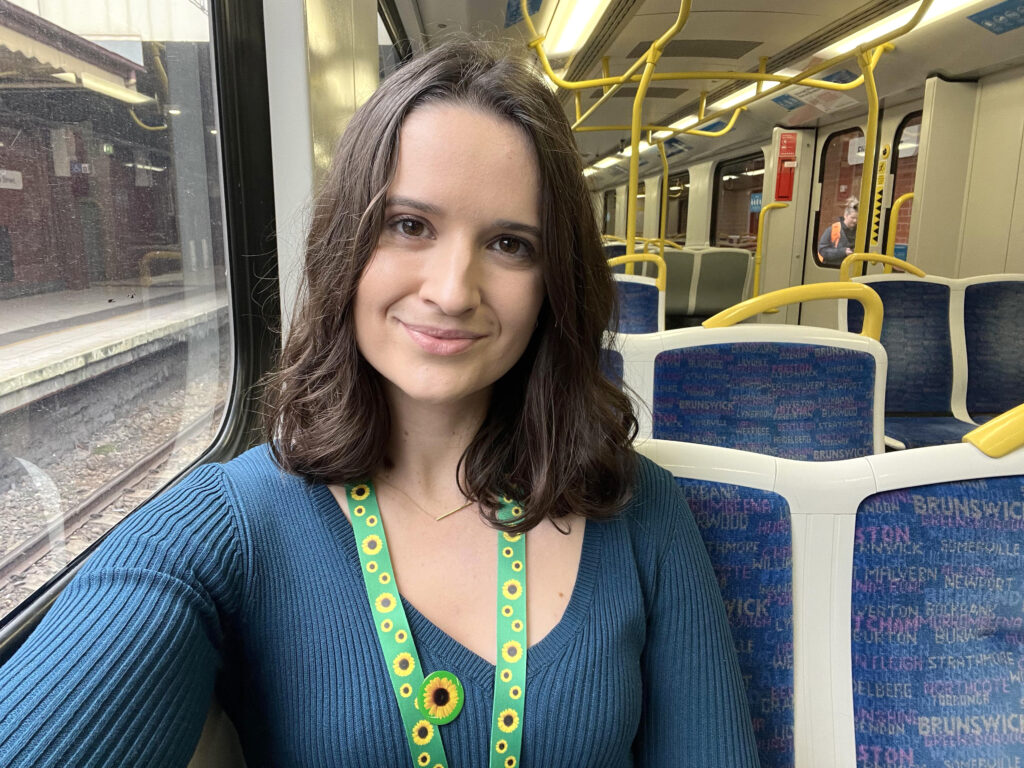 A young woman with short brown hair is sitting inside a train with yellow and blue seats in the background. She is smiling and wearing a bright green and yellow hidden disability sunflower lanyard and badge.