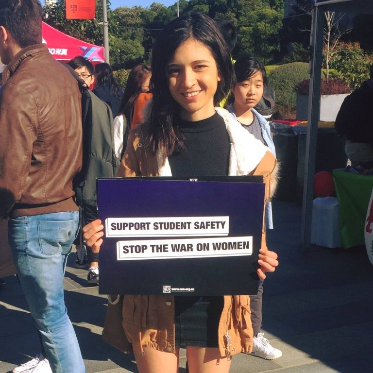 Photo of 23 year old Heidi at University of Sydney O’Week holding a purple sign with highlighted text reading: ‘Support Student Safety. Stop The War On Women.’
Heidi is wearing a black top and skirt and a brown coat. She has long dark brown hair and is smiling.