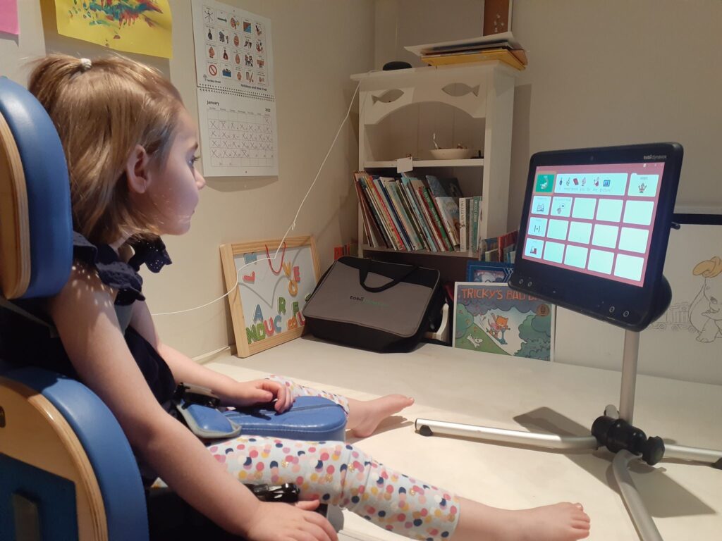 Izzy sitting in front of her Tobii Dynavox eye gaze computer, sitting on a stand on the desk. There are shelves, books, charts, a bag and pictures in the background. Izzy has white skin and shoulder length blonde hair, top half tied back.  She is wearing white leggings with coloured dots and a top with navy blue frills at the collar.