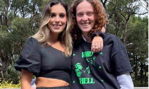 Two feminine featured people are looking at the camera smiling. My sister, on the left, has her arm around my shoulders. She’s wearing a black crop top and a long black flowing skirt. I’m wearing a black and green t-shirt and brown pin-striped pants. Behind us are tall gum trees