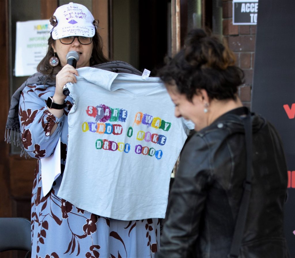 A photo of Hannah holding a microphone that she is speaking into, as well as a light grey shirt with coloured text that reads “Stare Away, I Know I Make Drool Cool.” Hannah  has fair skin and long dark brown hair, and is wearing a blue dress with a black floral pattern, glasses, earrings, a grey scarf, and a white cap with purple text. In the foreground is the blurred image of a person smiling with their back to the camera. They have fair skin, short dark brown hair, and are wearing a black leather jacket.