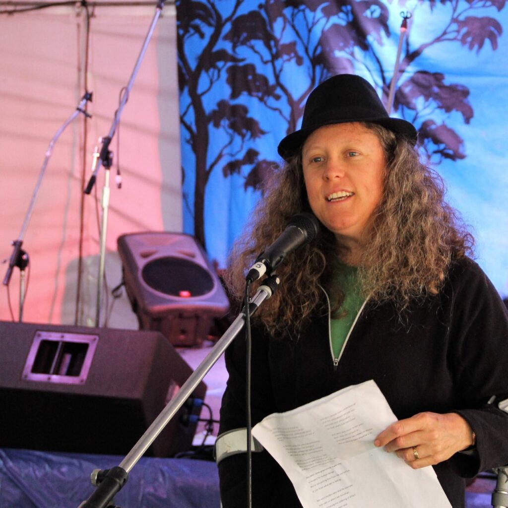 A photo of Jane speaking into a microphone on a stand. She has fair skin, long curly grey hair, and is wearing a black jacket, green t-shirt, and a black hat. She is holding speech notes, and behind her is a stage set up with amps, more microphones, and a blue and red background with the outline of black trees. 