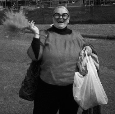 Laura, a middle aged white woman with blonde hair, standing on an oval. She is wearing a top and pants with whacky glasses and holding a large feather and white plastic bag in either hand.