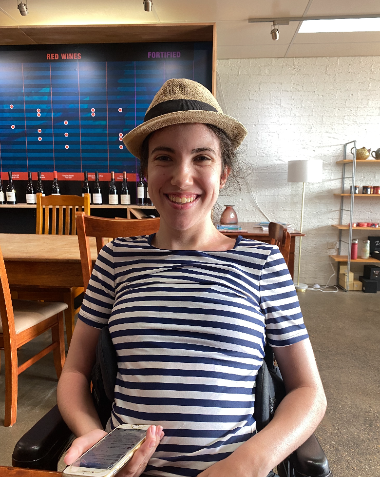 A photo of Hannah: a smiling woman with dark hair, wearing a hat, and sitting in a chair, phone in hand.