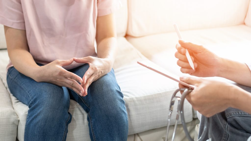  A doctor consulting a patient. A person with white skin from the shoulders down is pictured sitting on a white couch. They are wearing a light pink t-shirt and blue denim jeans, holding their hands together anxiously. Next to them are the hands of a doctor, holding a pen, notepad and stethoscope. The doctor has white skin and is wearing a white coat and jeans. 