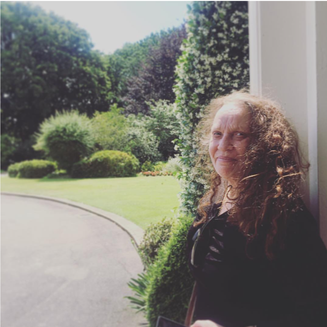 [Image Description: A photo of Cheryl, an Indigenous woman with pale skin and curly brown hair below her shoulders. She is standing sideways, looking at the camera with a closed mouth smile. She is wearing a black top, standing in front of a garden with bushes and trees in the background.]